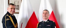 Pomorski komendant wojewódzki Państwowej Straży Pożarnej uściska dłoń funkcjonariuszowi wręczając mu dokument. Za nimi stoją flagi państwowe.