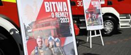 Na tle samochodów pożarniczych ustawione sztalugi z plakatem dotyczącym akcji : "Bitwa o remizy 2023". Na plakacie uwidoczniono: po lewej stronie na górze na jasnym tle znak wodny - godło RP. Po przekątnej od górnej prawej strony czerwony prześwitujący pas. na jego tle po prawej stronie na górze napis: "Bitwa o remizy 2023" Poniżej od lewej stronie kaskadowo ustawieni trzej strażacy przedstawieni od pasa. Wszyscy w ubraniach specjalnych koloru piaskowego, drugi i trzeci w hełmach koloru białego.
Na dalszym planie po prawej stronie przed samochodem pożarniczym ustawione drugie sztalugi z tym samym plakatem.