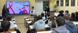 Na zdjęciu widać młodzież ze szkoły średniej z VI LO w Białymstoku podczas zajęć edukacyjnych, które prowadzą dwie dziewczyny i jeden chłopak. Klasa siedzi tyłem do obiektywu, a za plecami osób prowadzących widać tablicę multimedialną z czerwoną wstążeczką (symbolem solidarności z osobami HIV +) oraz napis, tytuł projektu „AIDStruction”.