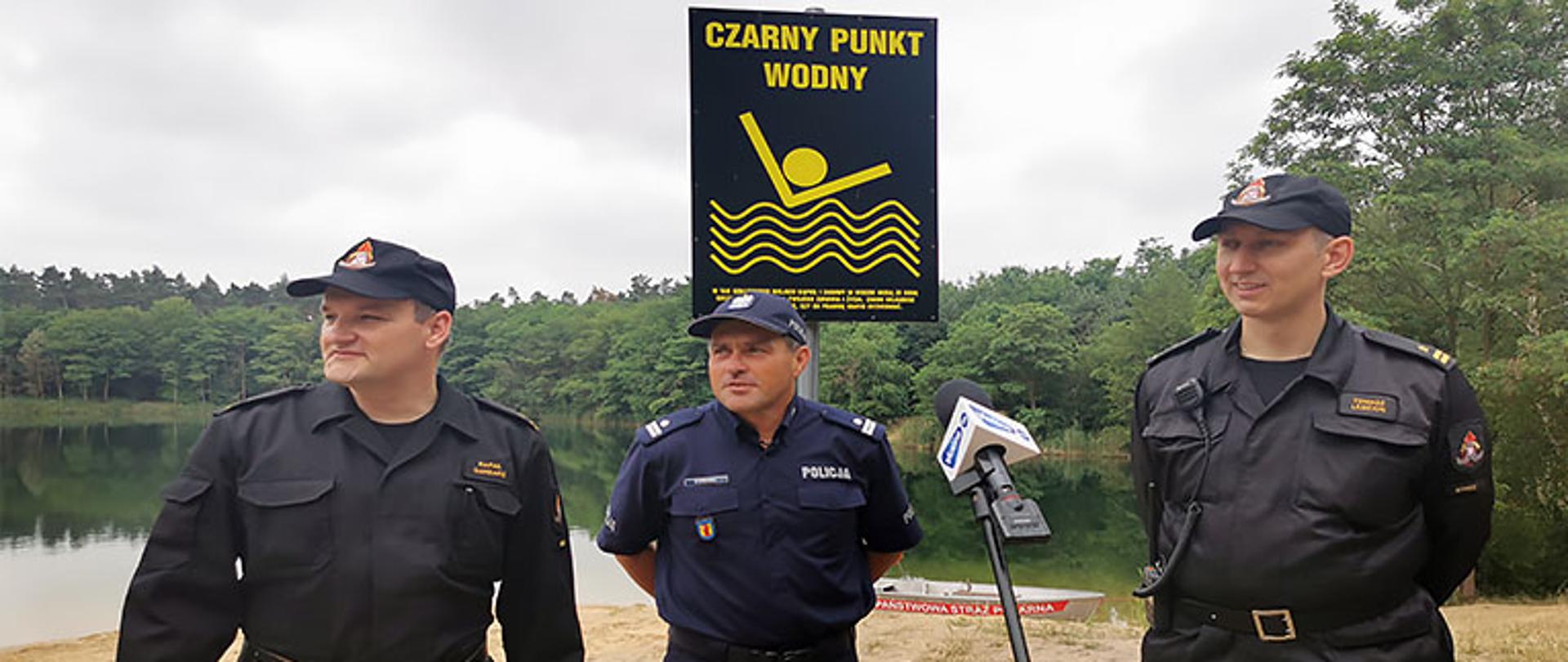 dwóch strażaków ubranych w ubranie koszarowe z czapką z daszkiem na głowie oraz jeden policjant w umundurowaniu z czapką z daszkiem na głowie udzielają wywiadu dla lokalnego radia, w tle zbiornik wodny oraz las który go okala, brzeg piaszczysty, lekkie zachmurzenie