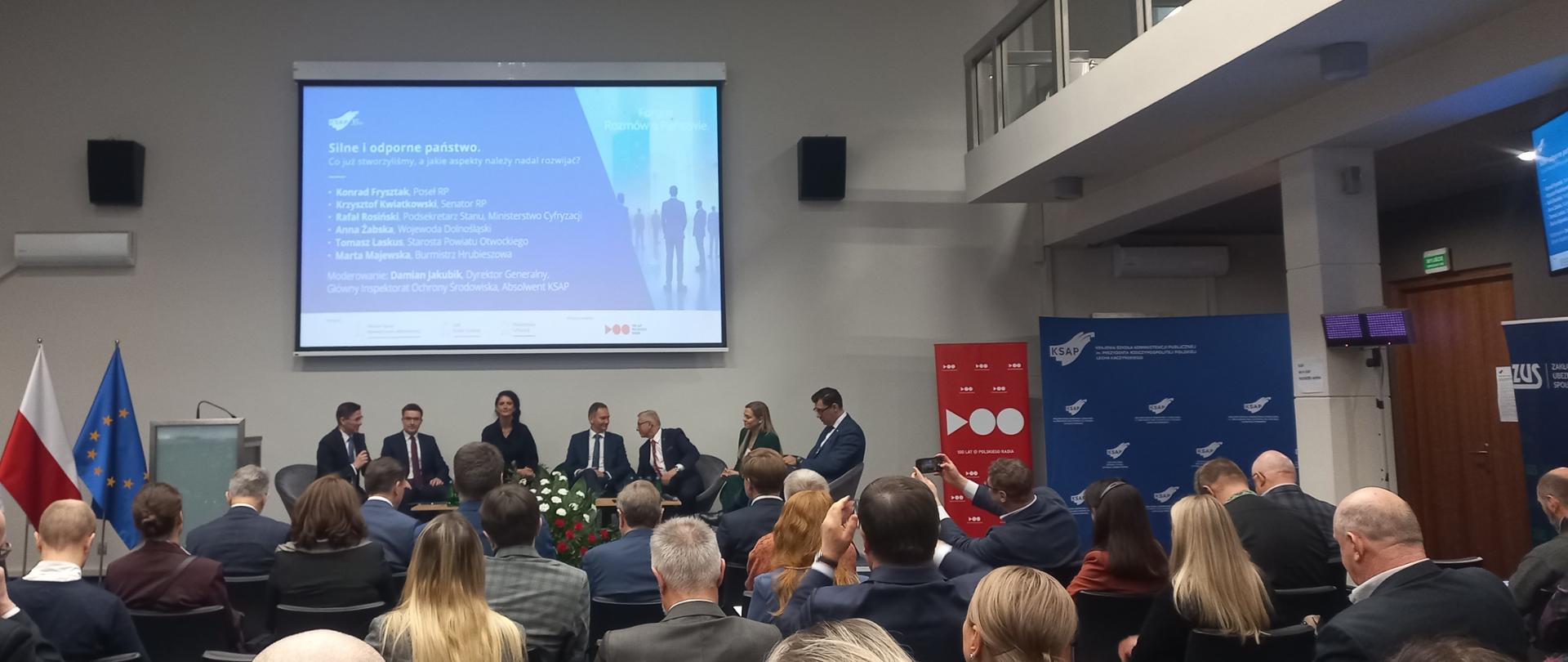 Duża grupa ludzi siedzi w sali, zwrócona w stronę prelegentów. 