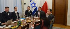 Przy podłużnym drewnianym stole siedzi kilka osób, u szczytu stołu minister Czarnek i mężczyzna w garniturze, za nimi flagi Polski i Izraela. Z lewej strony zasłonięte firanką okno.