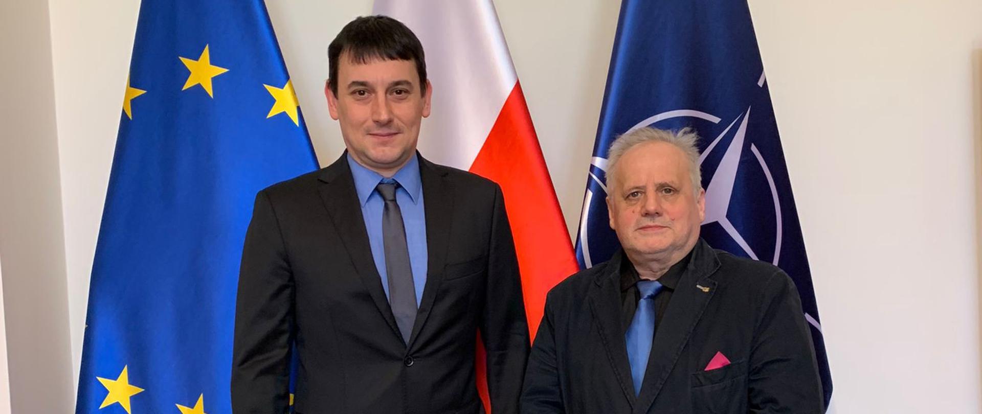 2 mężczyzn, w tym wicewojewoda, stoją obok siebie, pozują do zdjęcia na tle flag Polski i UE