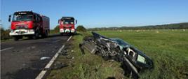 Fotografia przedstawia ujęcie wzdłuż pasa jezdni. Na jezdni przodem do fotografującego stoją dwa pojazdy straży pożarnej, po prawej stronie kadru w przydrożnym rowie znajduje się wrak rozbitego pojazdu osobowego w kolorze czarnym, pojazd ma widoczne zgniecenie w okolicy przedniego lewego koła, zniszczoną przednią szybę, widać ślady użycia hydraulicznych narzędzi ratowniczych.