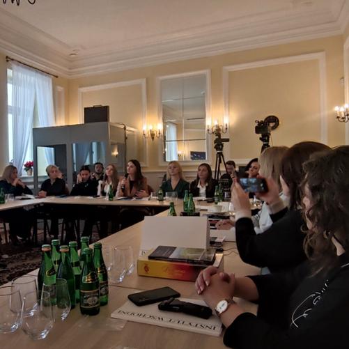 W sali konferencyjnej przy ustawionych w prostokąt stołach siedzą uczestniczki warsztatów i słuchają przemawiającej kobiety