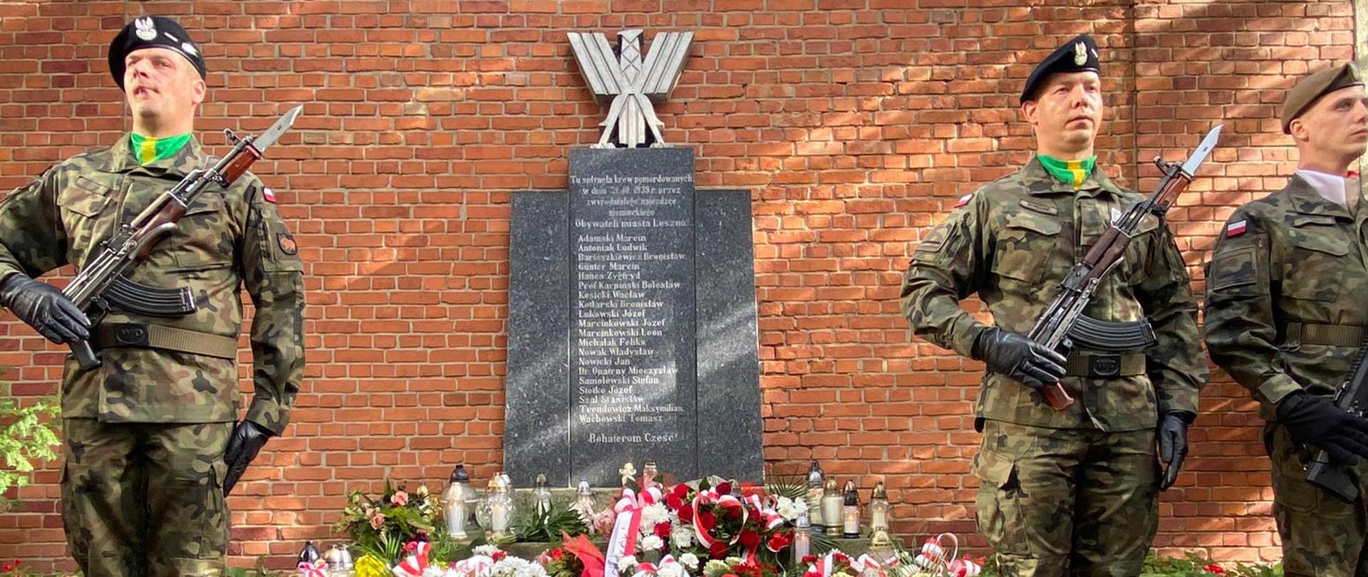 Żołnierze w mundurach z karabinami stoją na tle pomnika.
