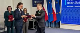 Prof. Dawid Sześciło odbiera akt powołania z rąk min. Jakuba Stefaniaka, sekretarza stanu, Zastępcy Szefa Kancelarii Prezesa Rady Ministrów; w tle flagi Polski i Unii Europejskiej