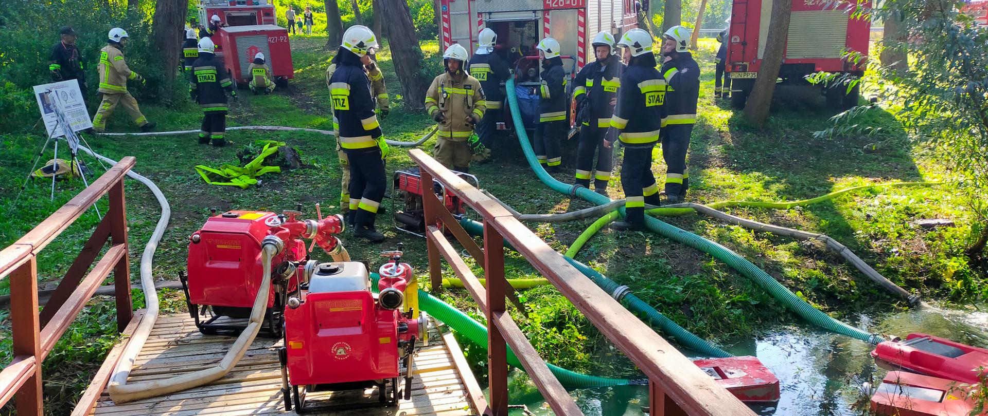 Punkt czerpania wody. Strażacy przy samochodach pożarniczych stojących tyłem . Na brzegu wąskiego strumienia rozwinięte węże. Na mostku stoją czerwone motopompy