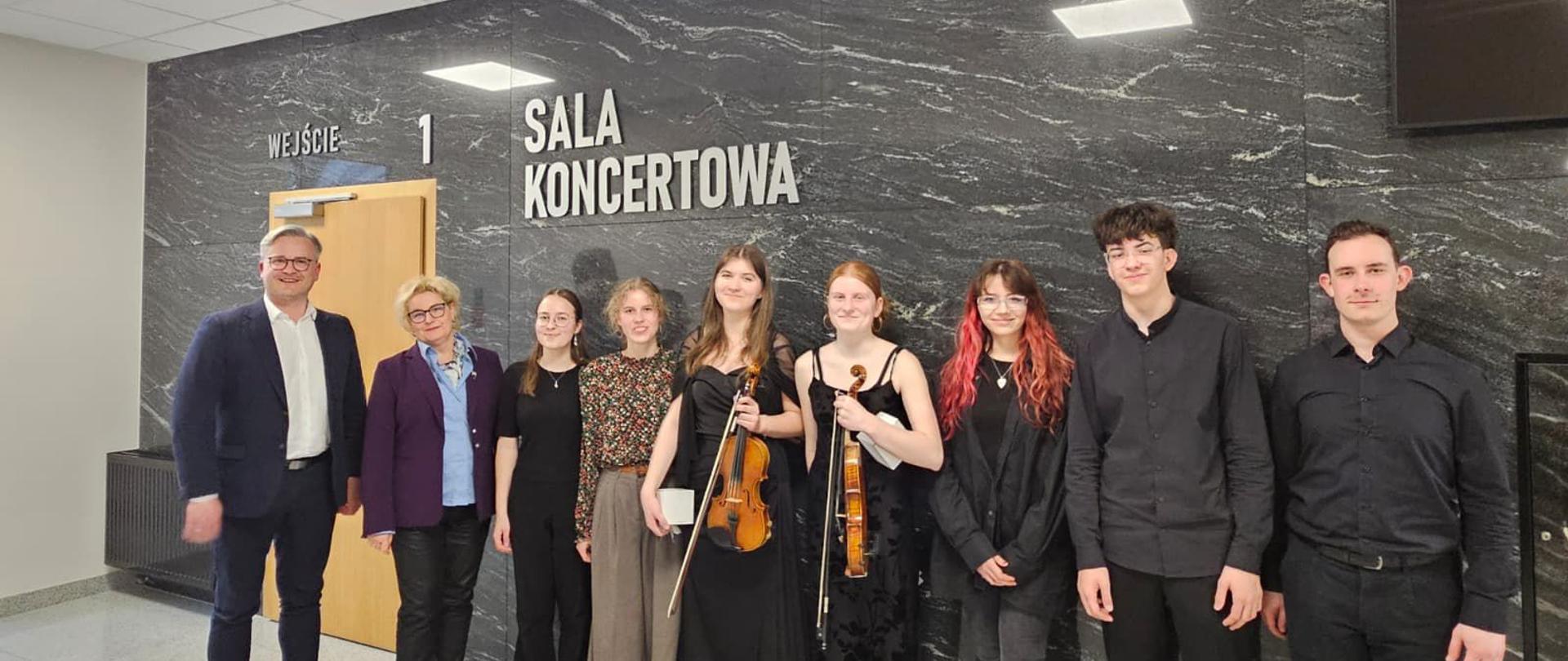 Na zdjęciu grupa młodych odświętnie ubranych artystów, którzy wystąpili podczas koncertu uczniów szkoły muzycznej II stopnia. Dwie osoby trzymają skrzypce . Zdjęcie zostało wykonane na tle czarnej kamiennej ściany. 