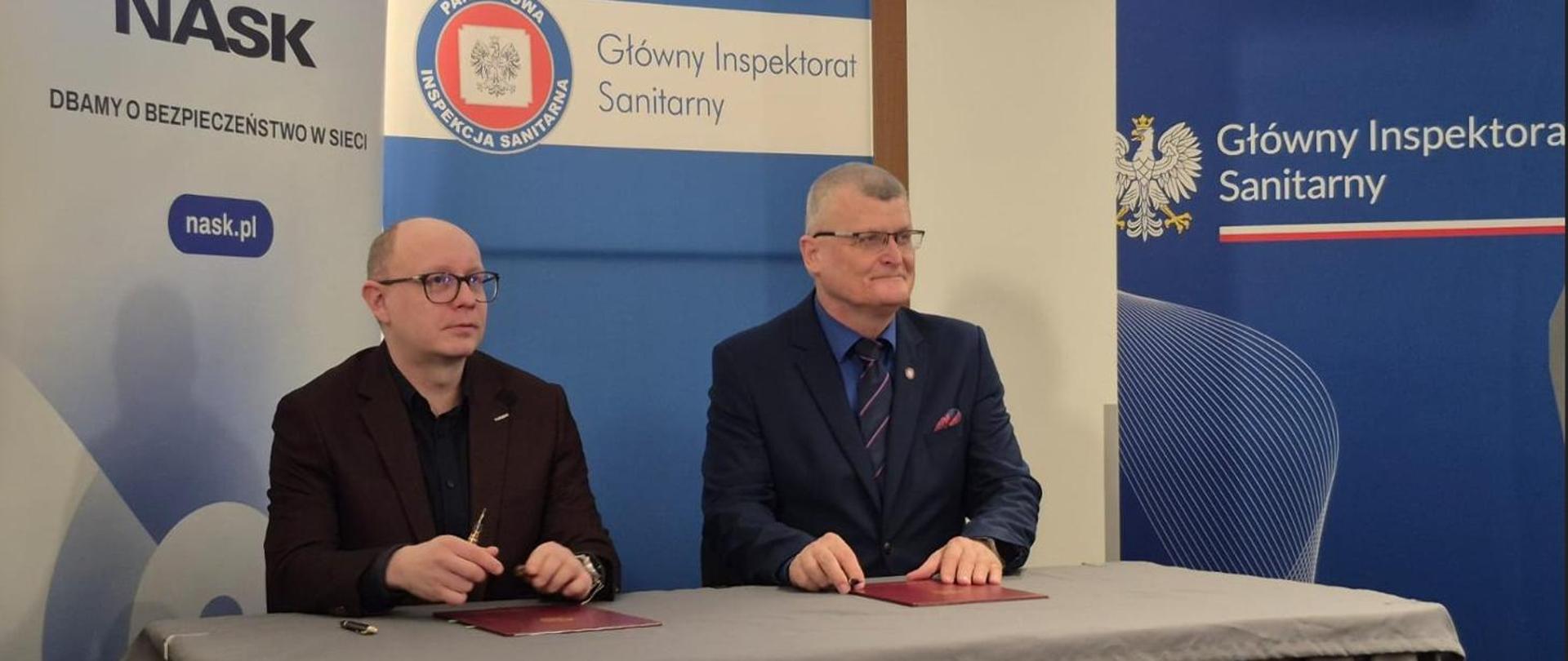 Na zdjęciu znajduje się dwóch mężczyzn w okularach siedzących za biurkiem