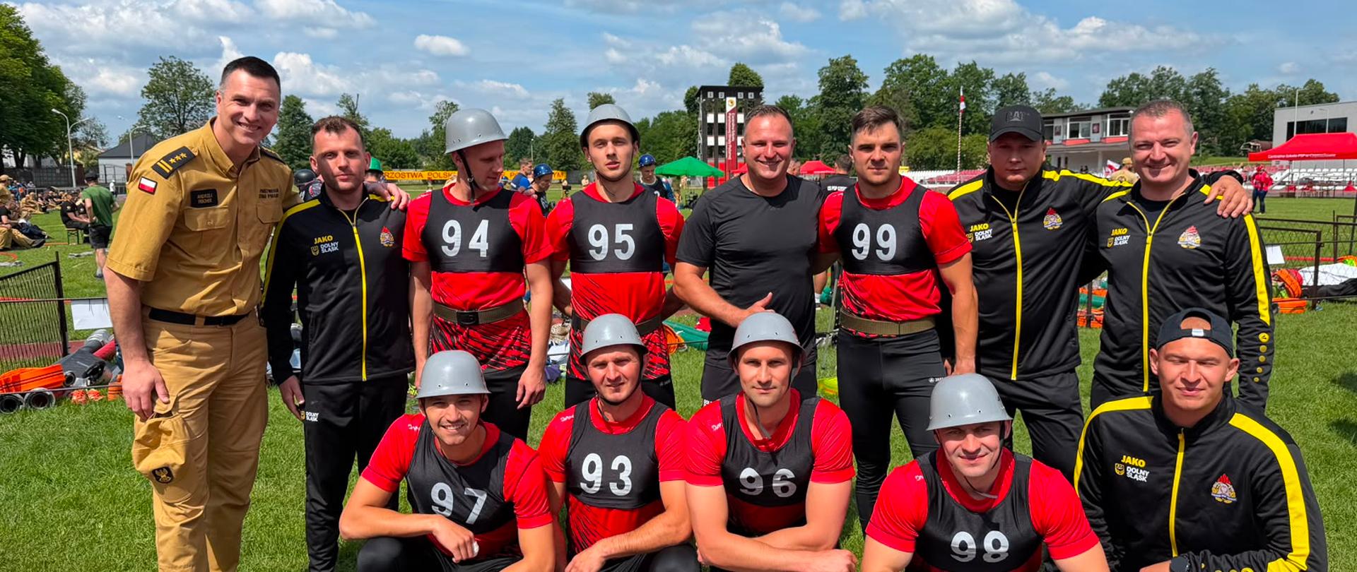 Grupowe zdjęcie zawodników i członków reprezentacji PSP Dolnego Śląska na stadionie sportowym podczas 42. Mistrzostw Polski w Sporcie Pożarniczym. Część osób ubrana jest w czerwono-czarne stroje startowe z numerami, a pozostali w czarno-żółte dresy oraz umundurowanie koszarowe. W tle widoczna jest murawa stadionu, elementy toru zawodów, trybuny oraz błękitne niebo z chmurami.