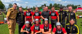 Grupowe zdjęcie zawodników i członków reprezentacji PSP Dolnego Śląska na stadionie sportowym podczas 42. Mistrzostw Polski w Sporcie Pożarniczym. Część osób ubrana jest w czerwono-czarne stroje startowe z numerami, a pozostali w czarno-żółte dresy oraz umundurowanie koszarowe. W tle widoczna jest murawa stadionu, elementy toru zawodów, trybuny oraz błękitne niebo z chmurami.