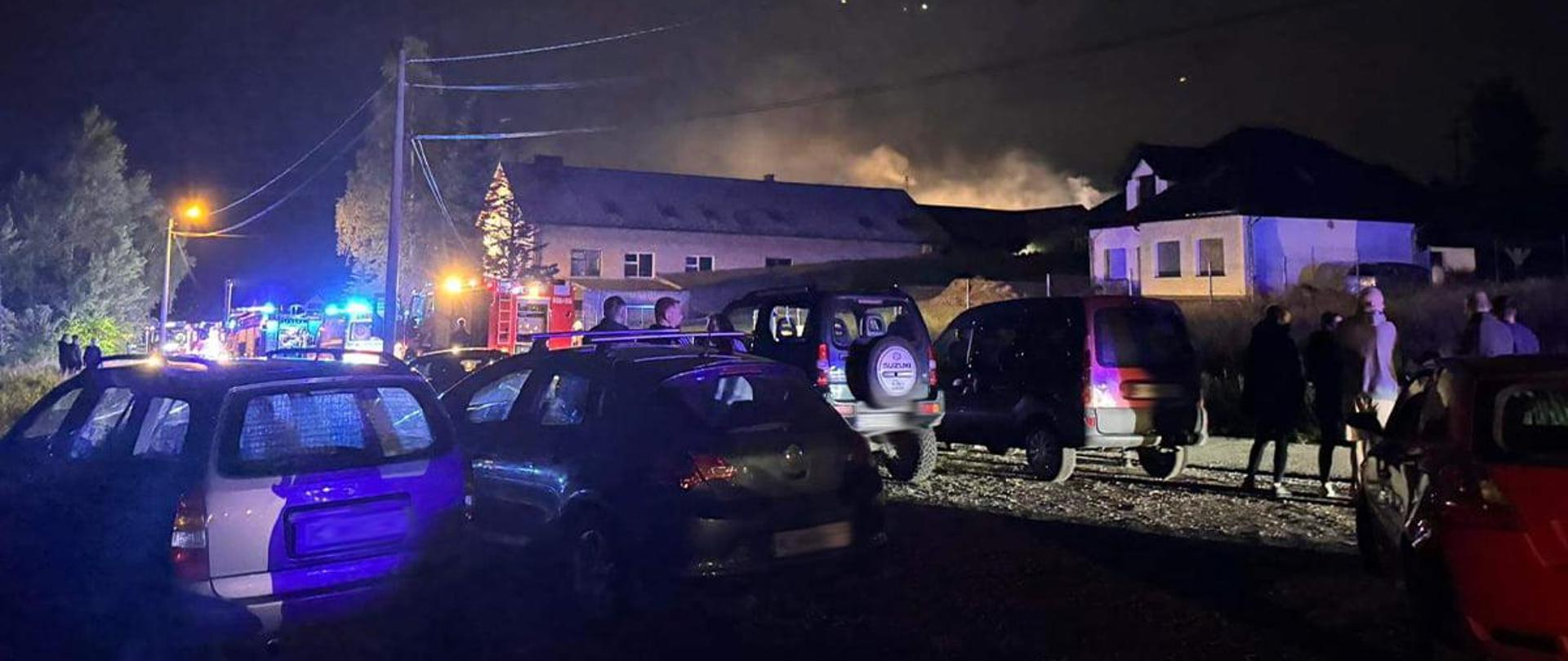 Widok z tyłu. W tle płonący budynek, z którego wydobywa się dym. Przed budynkiem, na drodze ustawione pojazdy pożarnicze jeden za drugim, na pierwszym planie samochody osobowe zaparkowane przy drodze oraz osoby obserwujące miejsce zdarzenia. 