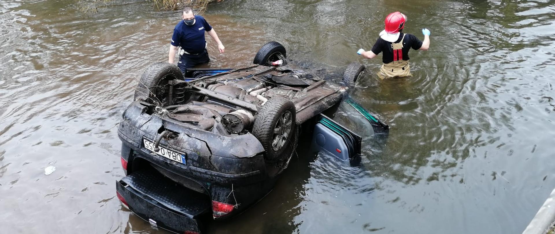 Samochód osobowy koloru czarnego znajduje się w korycie rzeki, widoczne otwarte 4 drzwi oraz otwarty bagażnik. Pojazd leży na dachu po lewej stronie stoi funkcjonariusz policji ubrany w mundur służbowy koloru granatowego mężczyzna w maseczce ochronnej - woda w której stoi sięga do połowy długości ud. po prawej stronie pojazdu stoi strażak skierowany twarzą do dowódcy znajdującego się na brzegu. Strażacy ubrani w ubrania specjalne typu nomeks.