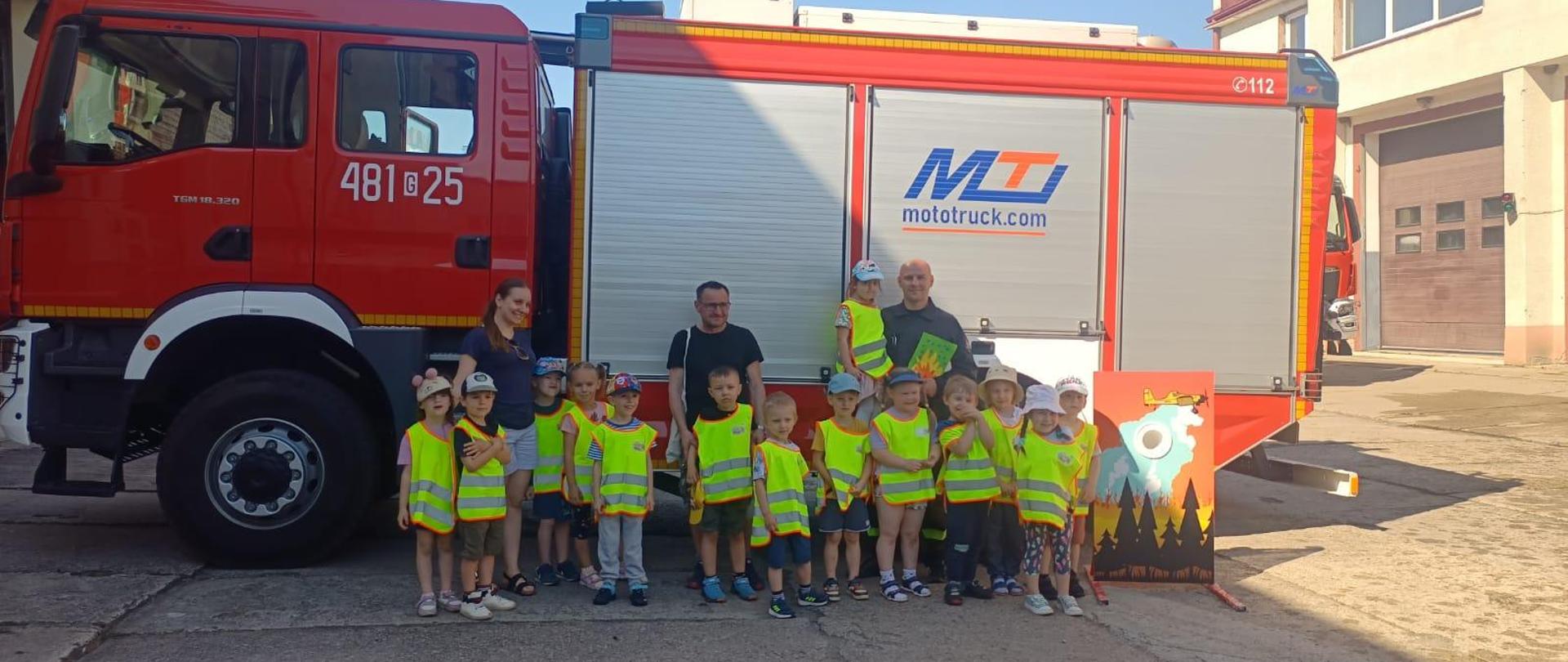 Dzieci z opiekunami i strażakiem na tle wozu strażackiego.