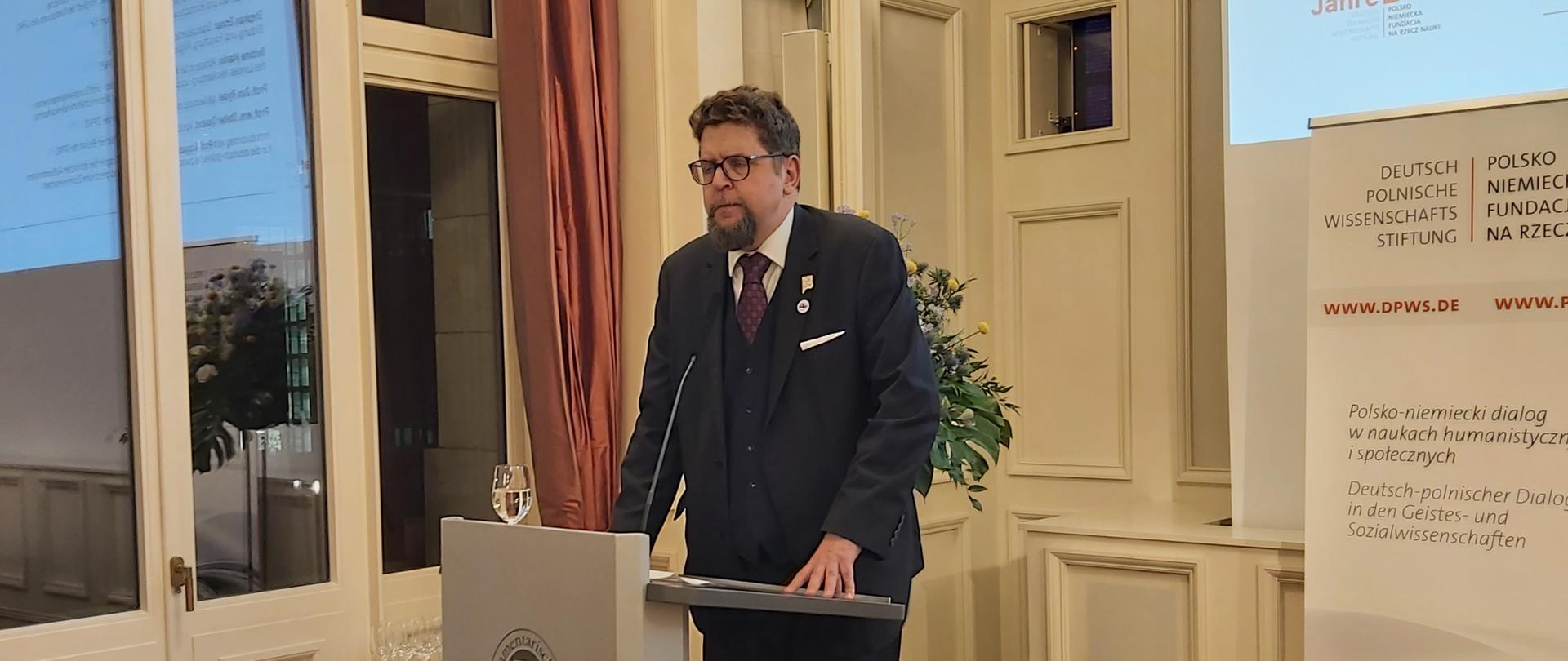 Wiceminister Szeptycki stoi za szarą mównicą i mówi do mikrofonu, obok niego czerwony fotel, z tyłu ściana, pod nią dwa wazony z kwiatami.