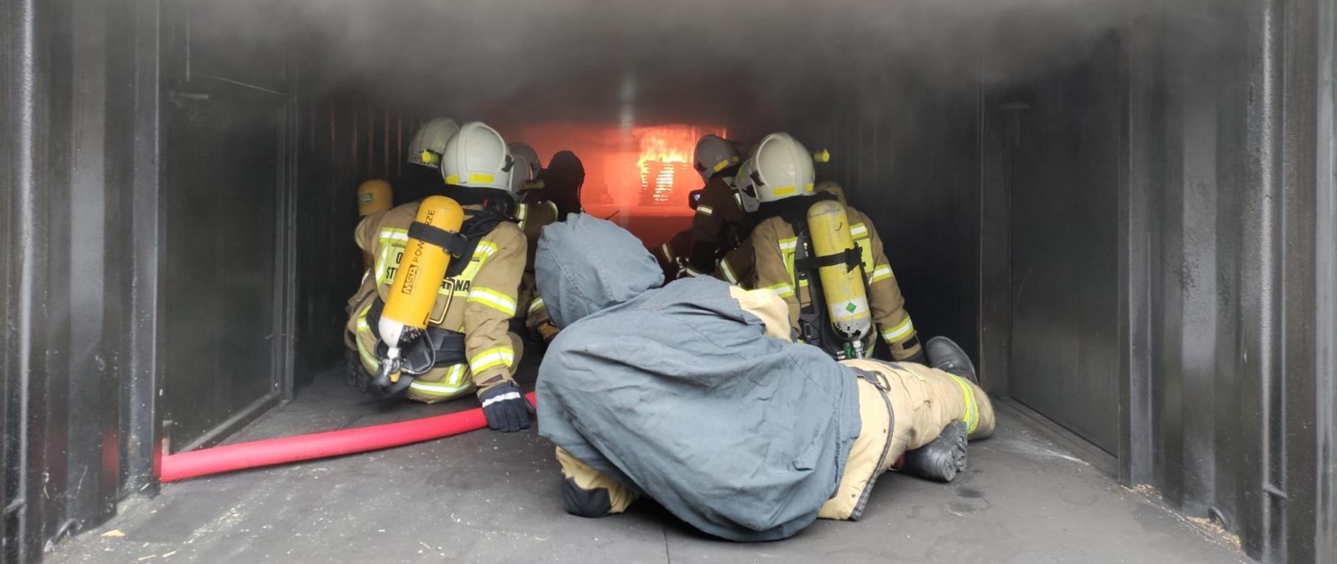 Strażacy OSP w ubraniu specjalnych w hełmach na głowie oraz w aparatach ochrony układu oddechowego uczestniczą w zajęciach w komorze rozgorzeniowej na poligonie w Komendzie Wojewódzkiej PSP w Opolu. 