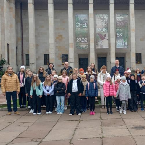 Zdjęcie zbiorowe 50-osobowej grupy dzieci, młodzieży, dorosłych. Wszyscy ustawieni są na tle budynku Muzeum Narodowe w Warszawie. Wszyscy są uśmiechnięci i ciepło ubrani.