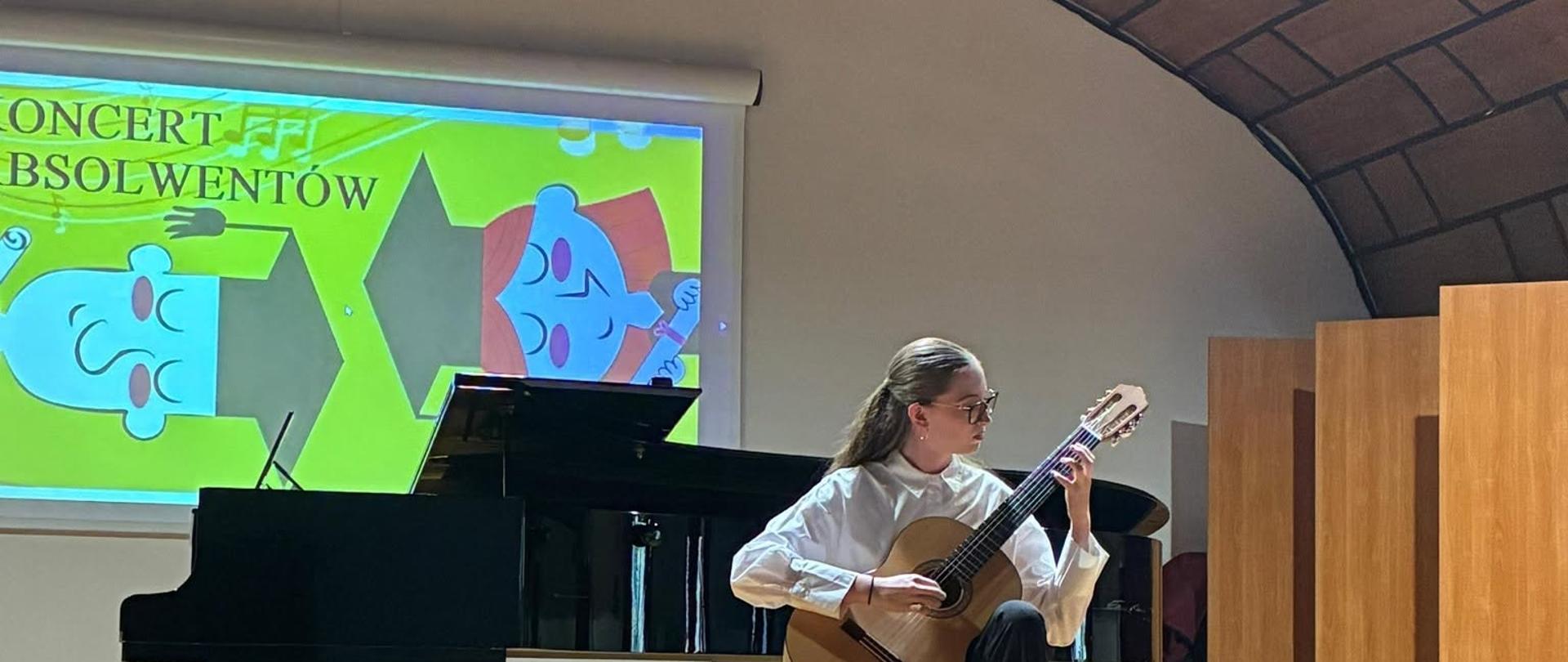 Młoda kobieta w okularach, białej koszuli i ciemnych spodniach siedzi na krześle na scenie i gra na gitarze klasycznej. Za nią stoi czarny fortepian skrzydłowy. W tle widać ekran z jasną grafiką i napisem „KONCERT ABSOLWENTÓW”. Scena ma drewnianą podłogę, a sufit sali jest łukowaty, pokryty kasetonami.