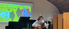 Młoda kobieta w okularach, białej koszuli i ciemnych spodniach siedzi na krześle na scenie i gra na gitarze klasycznej. Za nią stoi czarny fortepian skrzydłowy. W tle widać ekran z jasną grafiką i napisem „KONCERT ABSOLWENTÓW”. Scena ma drewnianą podłogę, a sufit sali jest łukowaty, pokryty kasetonami.