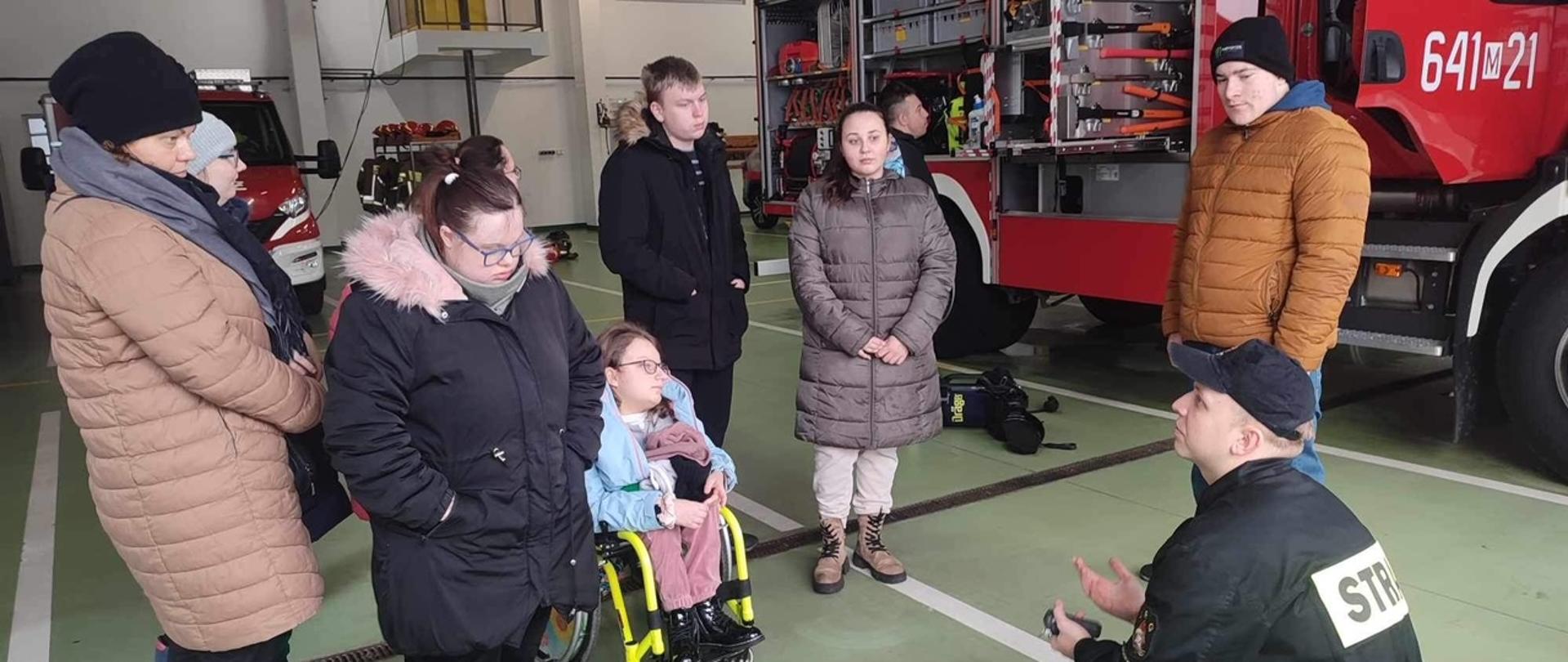 Wycieczka w ramach akcji „Odwiedź strażnicę i salę edukacyjną typu „Ognik” w KP PSP w Sokołowie Podlaskim 22.02.2024 r. - na zdjęciu młodzi adepci pożarnictwa zapoznają się z pojazdami i wyposażeniem znajdującym się w jednostce, przed czerwonym samochodem strażackim.