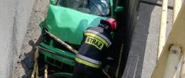 Zdjęcie przedstawia zielony samochód który wpadł pomiędzy mur oporowy budynku a jezdnie na głębokość ok 2 metrów