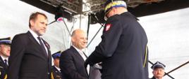 Dwóch mężczyzn stoi na scenie. Strażak wręcza jednemu z nich dyplom. Za nimi stoją strażacy