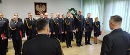 Zdjęcie przedstawia funkcjonariuszy podczas uroczystości mianowania na wyższe stanowiska służbowe. Strażacy stojący w dwuszeregu, znajdują się w świetlicy Komendy Powiatowej Państwowej Straży Pożarnej w Lwówku Śląskim. Przed strażakami znajduje się Komendant Powiatowy PSP w Lwówku Śląskim oraz Dowódca Jednostki Ratowniczo-Gaśniczej PSP w Lwówku Śląskim.