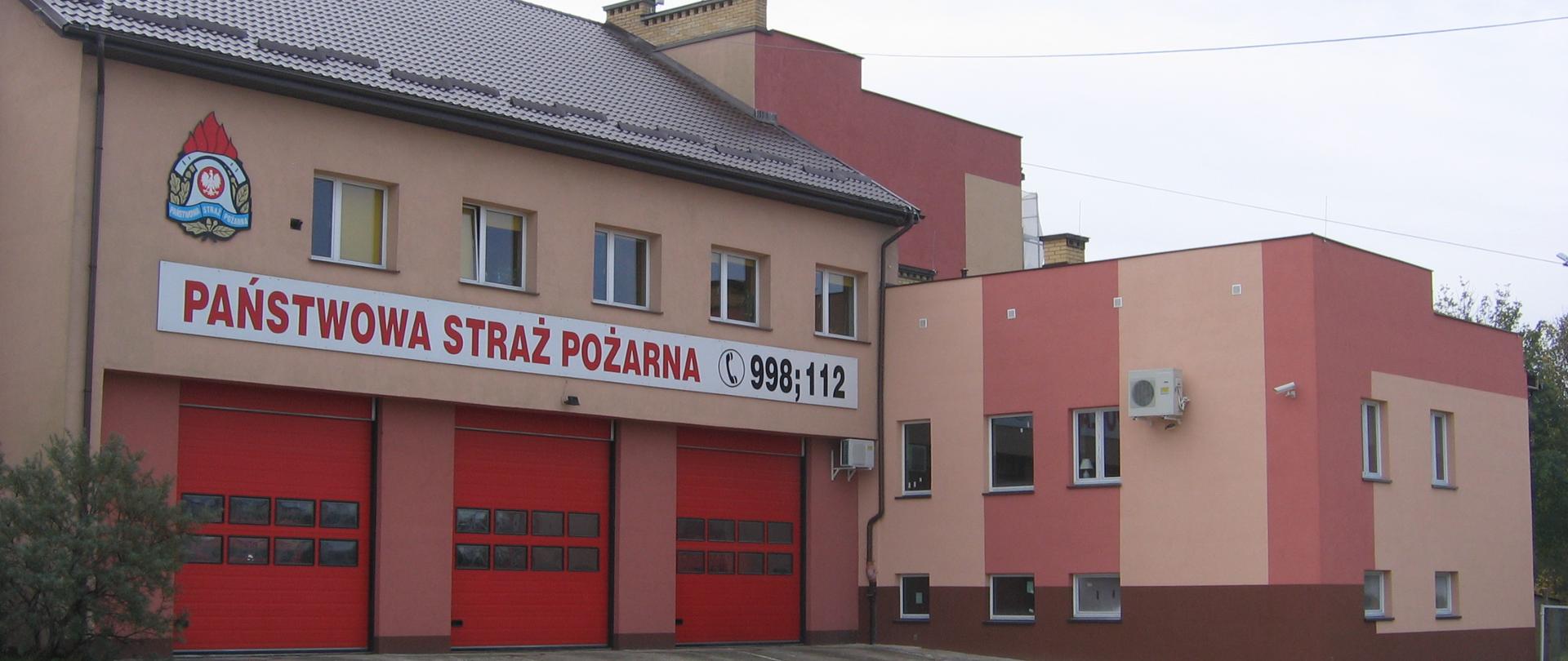Budynek Komendy Powiatowej PSP w Hajnówce z trzema czerwonymi bramami garażowymi. Elewacja w odcieniach koloru różowego. Nad bramami napis Państwowa Straż Pożarna 998, 112.