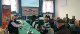 Na zdjęciu widać 2 strażaków PSP oraz 12 osób w normalnych ubraniach siedzących przy dwóch stołach w czasie spotkania