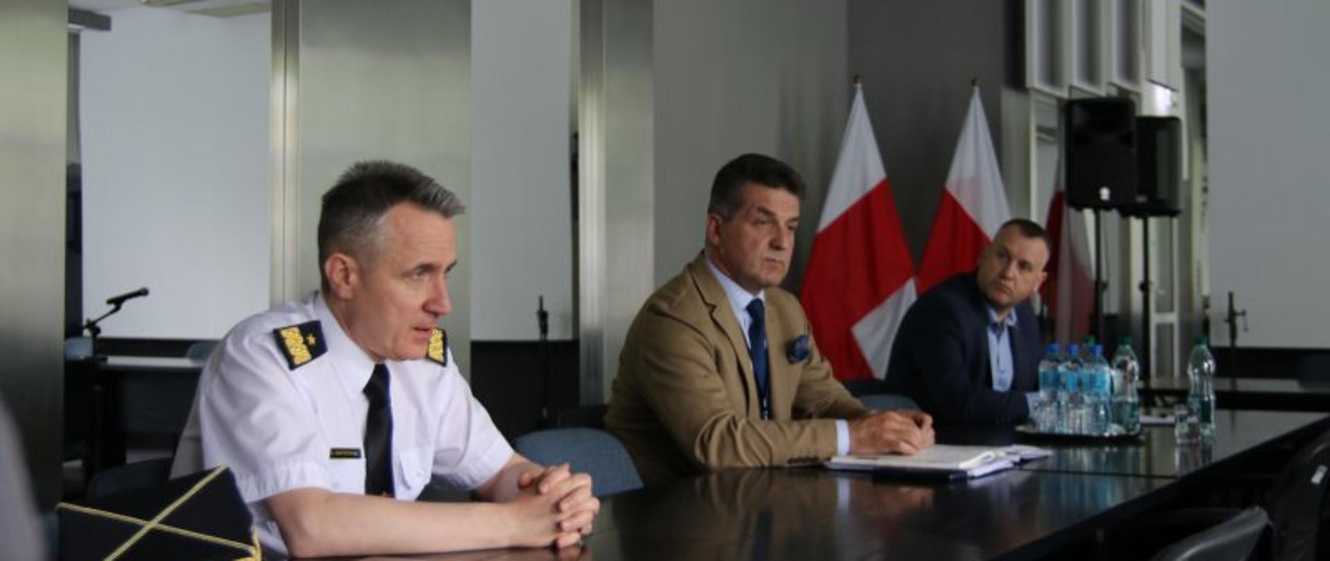 Spotkanie Komendanta Głównego PSP z Prezydium Zarządu Krajowego ZZS „Florian” – na zdjęciu uczestnicy spotkania dyskutują przy stole. W tle dwie flagi biało-czerwone.