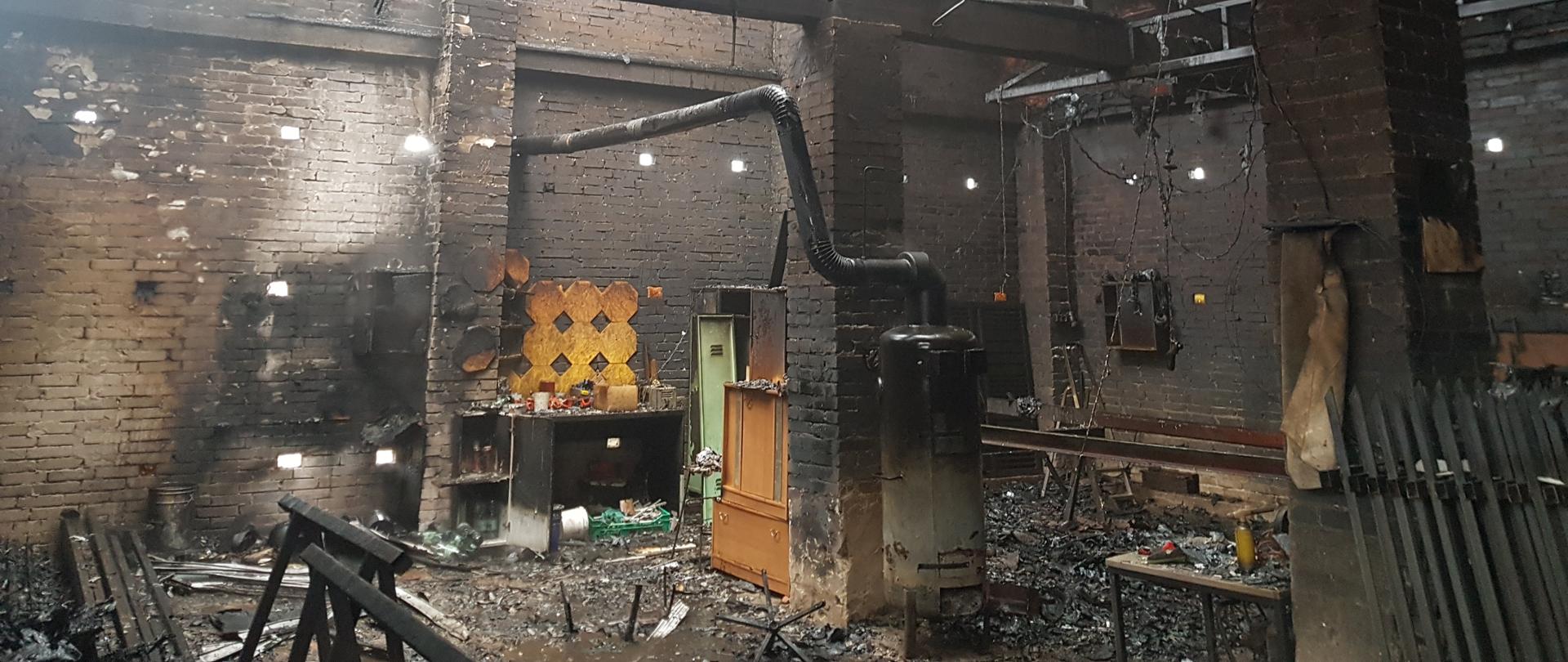 Zdjęcie przedstawia wnętrze budynku gospodarczego po pożarze. Widoczne są ściany, konstrukcja dachu oraz posadzka. Po prawej stronie zdjęcia widoczne są metalowe przęsła ogrodzenia, które zostały oparte o słup wybudowany z białej cegły. Po prawej stronie widoczne są także długie, stalowe belki oraz aluminiowa, rozkładana drabinka. Obok słupa znajduje się stolik, na którym znajdują się małe przedmioty. Na środku zdjęcia znajduje się kolejny słup, obok którego znajduje się blaszany, czarno – szary piec, od którego poprowadzona jest instalacja odprowadzania spalin w postaci metalowej rury - prowadzi spaliny przez otwór w ścianie, na zewnątrz budynku. Na słupach wykonanych z cegły, spoczywają masywne belki – stanowią konstrukcje budynku. Można zauważyć dużo zwisających z góry przewodów elektrycznych. Po drugiej stronie słupa znajdują się zniszczone
i okopcone dwie szafki: jedna stalowa, druga jasna z płyty fornirowanej. Pod ścianą znajduje się stolik. Po lewej stronie zdjęcia znajdują się stalowe konstrukcje i podłużne elementy. W pokryciu dachu znajduje się wiele otworów, przez które wpada światło dzienne. Na posadce leży dużo okopconych i spalonych przedmiotów. Ściany budynku oraz słupy są okopcone na czarno. 