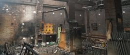 Zdjęcie przedstawia wnętrze budynku gospodarczego po pożarze. Widoczne są ściany, konstrukcja dachu oraz posadzka. Po prawej stronie zdjęcia widoczne są metalowe przęsła ogrodzenia, które zostały oparte o słup wybudowany z białej cegły. Po prawej stronie widoczne są także długie, stalowe belki oraz aluminiowa, rozkładana drabinka. Obok słupa znajduje się stolik, na którym znajdują się małe przedmioty. Na środku zdjęcia znajduje się kolejny słup, obok którego znajduje się blaszany, czarno – szary piec, od którego poprowadzona jest instalacja odprowadzania spalin w postaci metalowej rury - prowadzi spaliny przez otwór w ścianie, na zewnątrz budynku. Na słupach wykonanych z cegły, spoczywają masywne belki – stanowią konstrukcje budynku. Można zauważyć dużo zwisających z góry przewodów elektrycznych. Po drugiej stronie słupa znajdują się zniszczone
i okopcone dwie szafki: jedna stalowa, druga jasna z płyty fornirowanej. Pod ścianą znajduje się stolik. Po lewej stronie zdjęcia znajdują się stalowe konstrukcje i podłużne elementy. W pokryciu dachu znajduje się wiele otworów, przez które wpada światło dzienne. Na posadce leży dużo okopconych i spalonych przedmiotów. Ściany budynku oraz słupy są okopcone na czarno. 