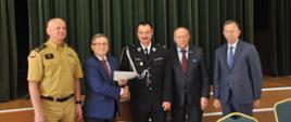 Na zdjęciu stoi dwóch strażaków jeden w mundurze PSP, drugi w mundurze OSP oraz trzech mężczyzn w garniturach (jeden z nich trzyma w ręce teczkę). Strażak OSP oraz jeden z mężczyzn podają sobie ręce. Z tyłu zielona kotara.