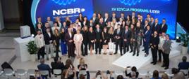 Zdjęcie od tyłu sali z góry, na sali na podwyższeniu z przodu pod wielkim niebieskim banerem z napisem NCBR - XV edycja Programu LIDER stoi grupa kilkudziesięciu osób, przed nimi ustawione w rzędy białe krzesła przed którymi stoją ludzie robiący zdjęcia.