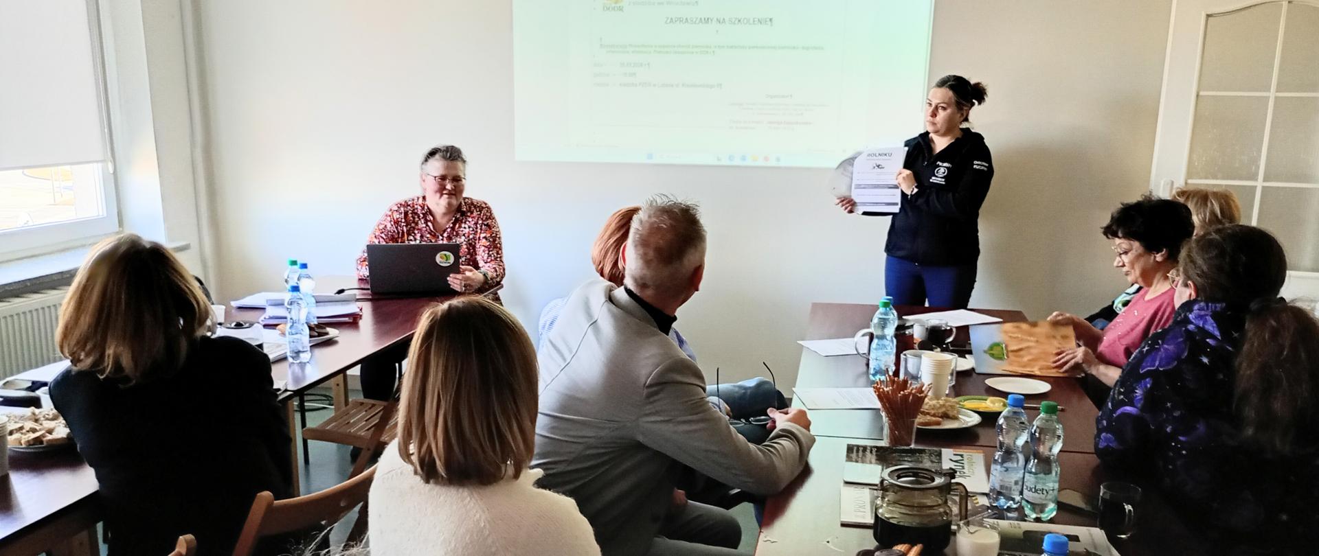 Grupa osób uczestniczy w szkoleniu w sali konferencyjnej. Uczestnicy siedzą przy dużym stole z notatkami, wodą i przekąskami. Z przodu jedna z prowadzących prezentuje dokument, a druga obsługuje laptop obok wyświetlanej na ścianie prezentacji.