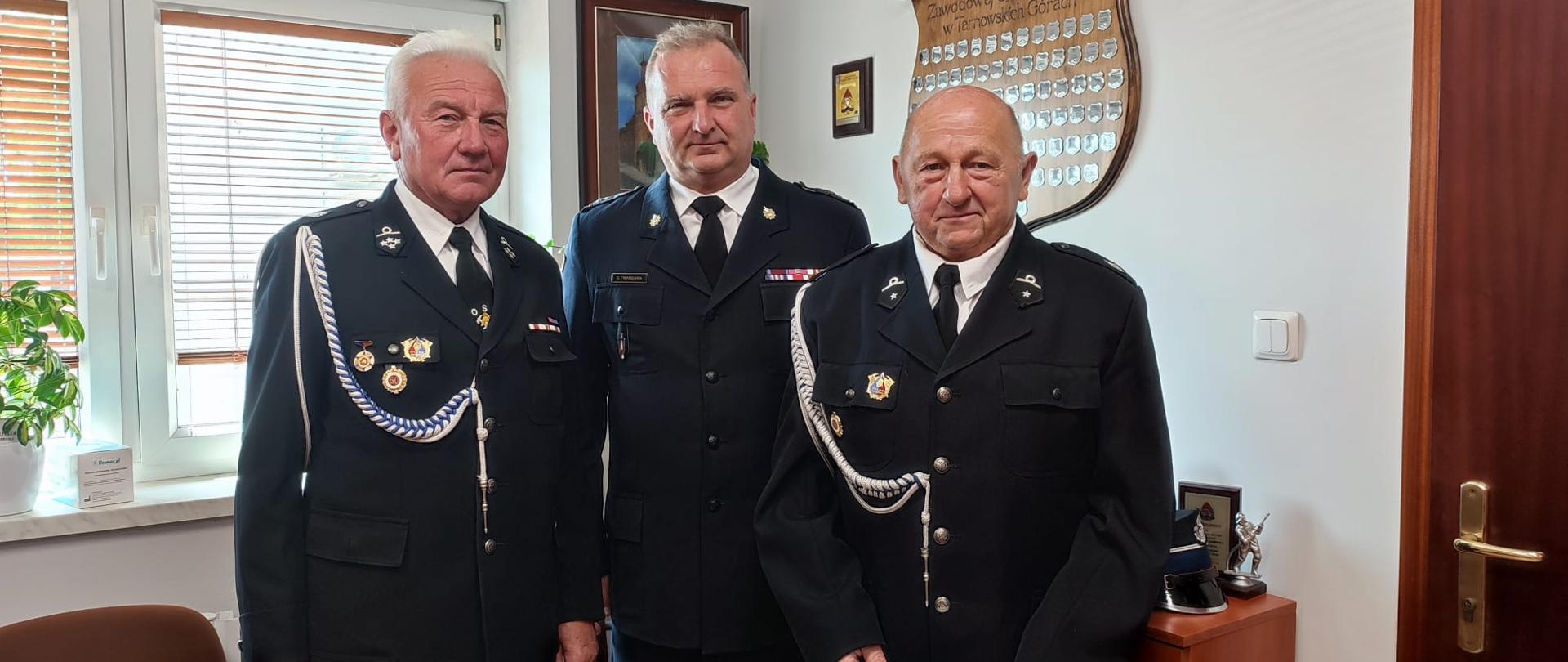 Trzech strażaków w umundurowaniu stoi w gabinecie. W środku Zastępca Komendanta Powiatowego PSP 