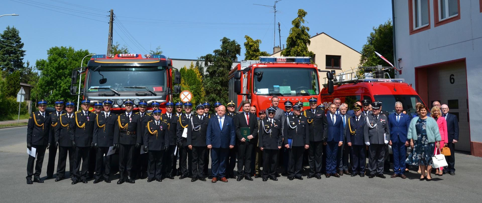 strażacy druhowie i inni uczestnicy uroczystości pozują do zdjęcia na tle samochodów pożarniczych z prawej strony widać bramę garażową z nr 6