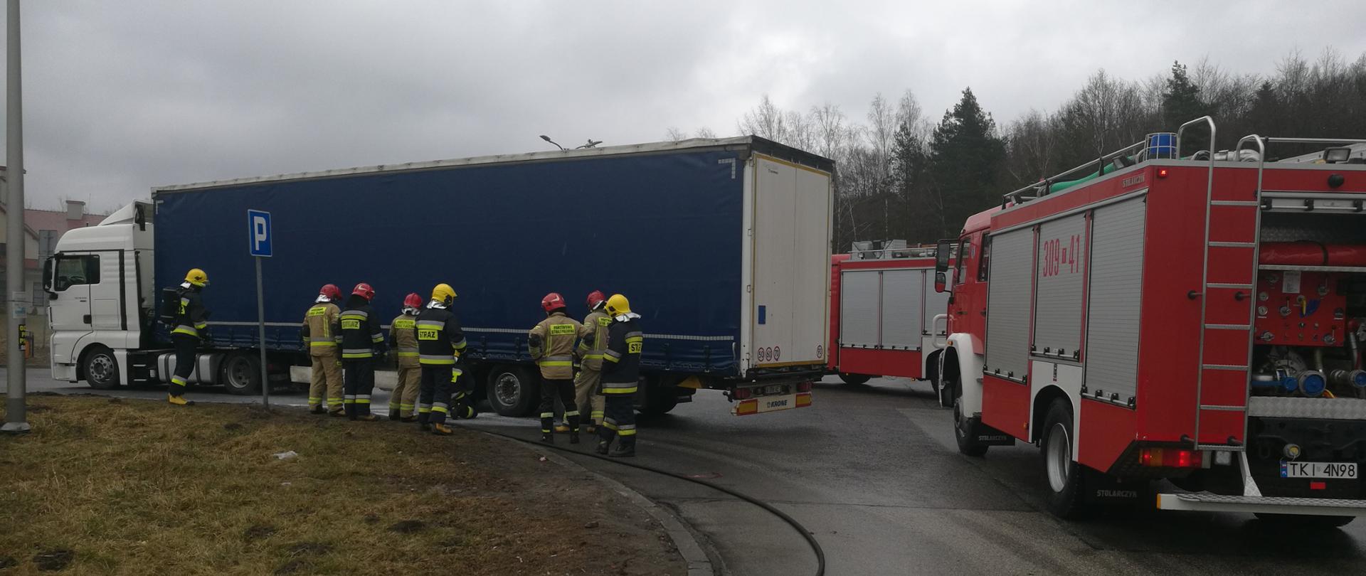 Zdjęcie przedstawia strażaków stojących przy naczepie samochodu ciężarowego. Na pierwszym planie widać rozwiniętą linię gaśniczą - obok stoi samochód Straży Pożarnej.