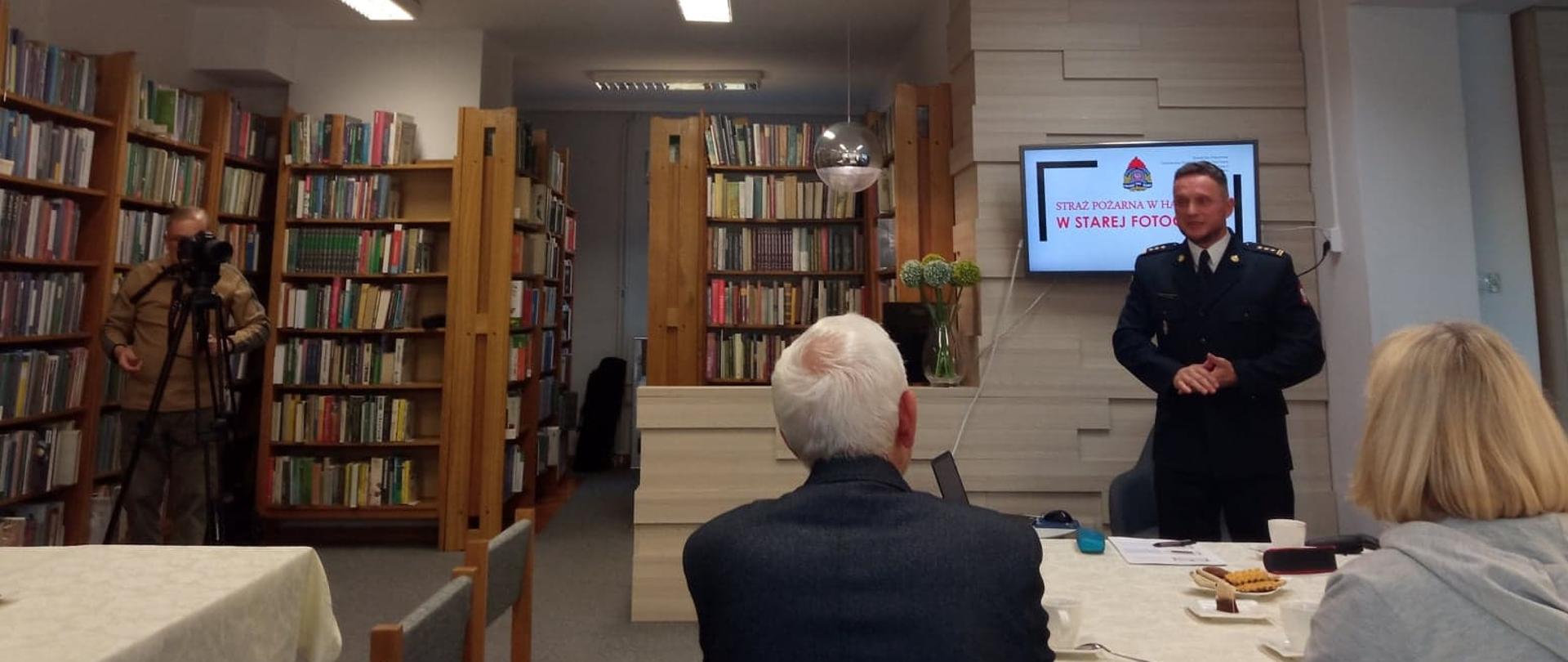 W pomieszczeniu czytelni stoi funkcjonariusz PSP ubrany w mundur. Przy stoliku siedzą dwie osoby słuchające wystąpienia strażaka.