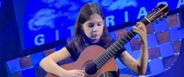 Na scenie zasiada młoda dziewczyna. W skupieniu gra na gitarze.
