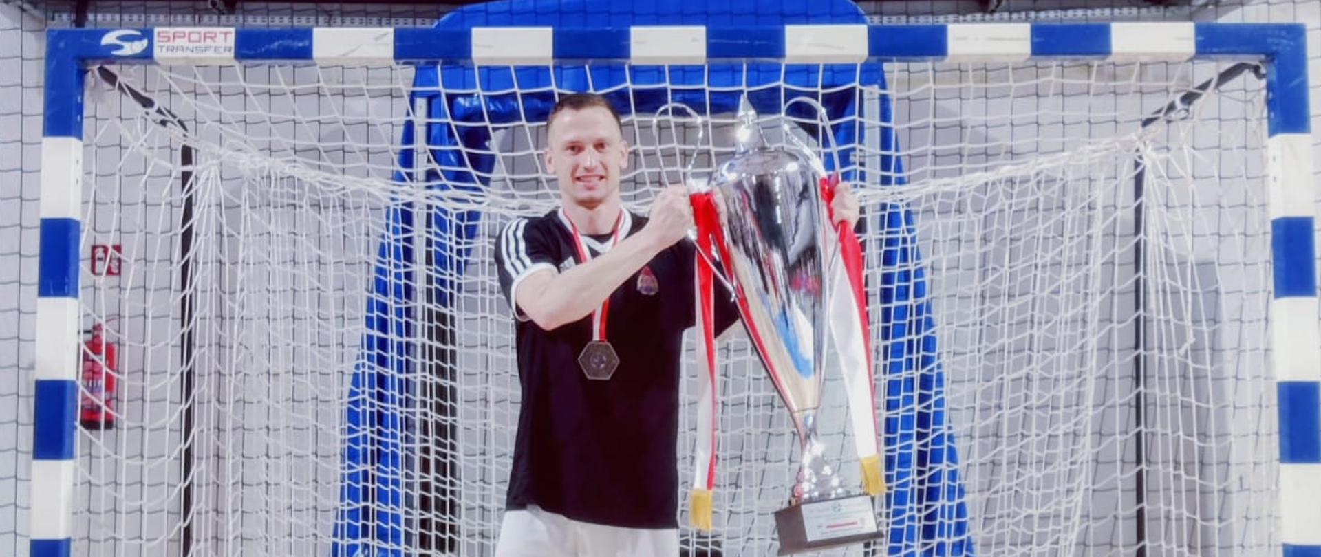 Na pierwszym planie zdjęcia widoczny jest mężczyzna ubrany w białą sportową koszulę i spodenki, na szyi ma złoty medal natomiast w ręku trzyma puchar. Znajduje się na hali sportowej.