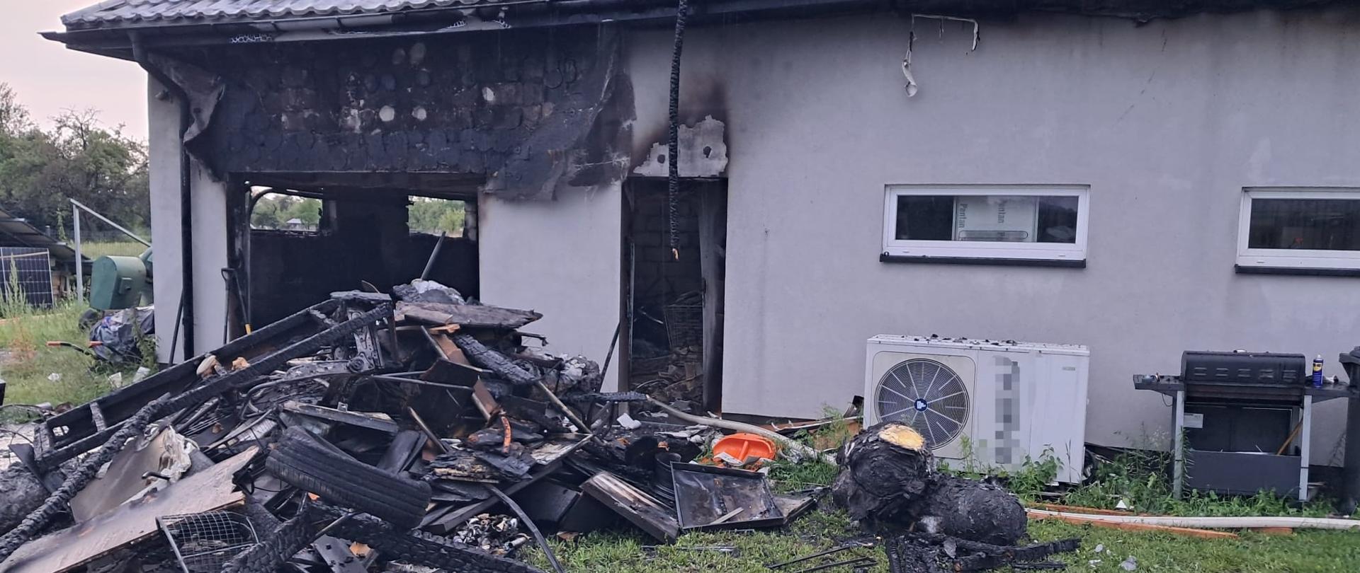 Spalony budynek gospodarczy z zapadniętym dachem i czarnymi ścianami sąsiaduje z zielonym żywopłotem i domem, a obok leżą stosy zwęglonych belek, opona i zniszczone urządzenia elektryczne.