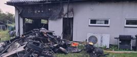 Spalony budynek gospodarczy z zapadniętym dachem i czarnymi ścianami sąsiaduje z zielonym żywopłotem i domem, a obok leżą stosy zwęglonych belek, opona i zniszczone urządzenia elektryczne.