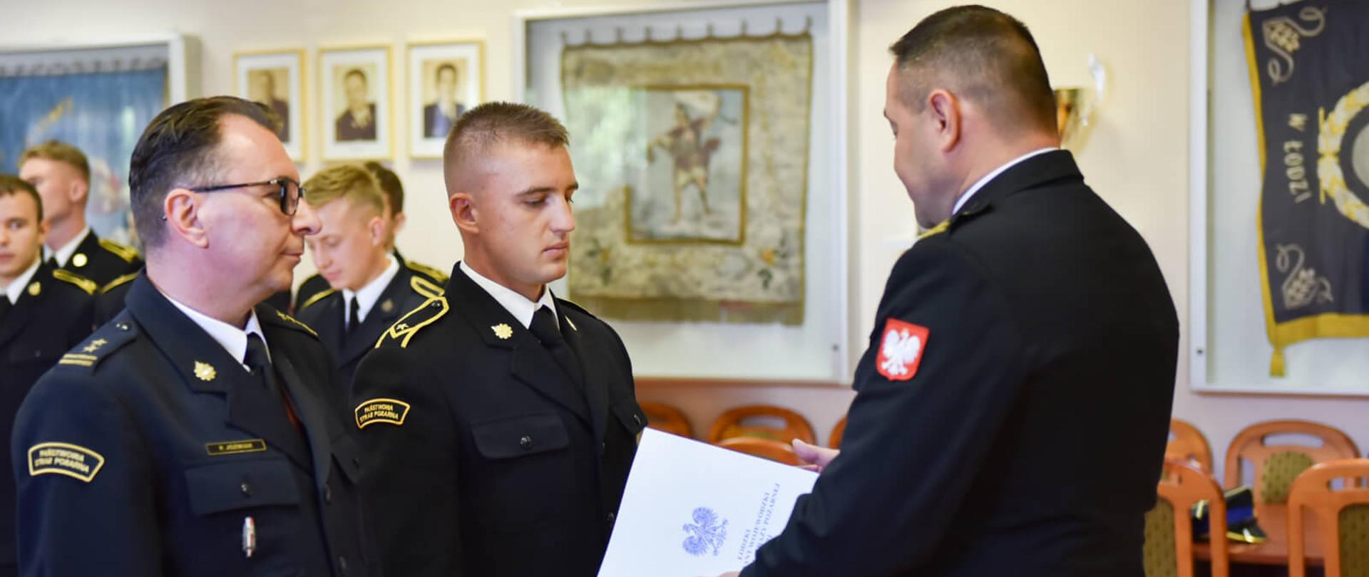 Na pierwszym planie zdjęcia widać trzech strażaków. Dwóch stoi obok siebie. Naprzeciwko stoi trzeci, który trzyma w ręku białą teczkę. Wszyscy ubrani są w ciemne stroje. W tle widać innych strażaków. Uroczystość odbywa się w pomieszczeniu, w którym zapalone jest światło.