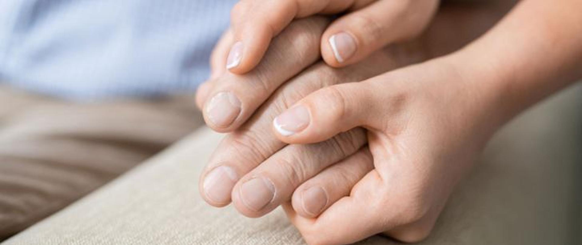 Dwie dłonie młodszej osoby trzymające dłoń starszej osoby