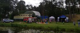 Dzieci nad wodą uczestniczą w zajęciach organizowanych przez funkcjonariuszy. W tle samochód strażacki i policyjny.
