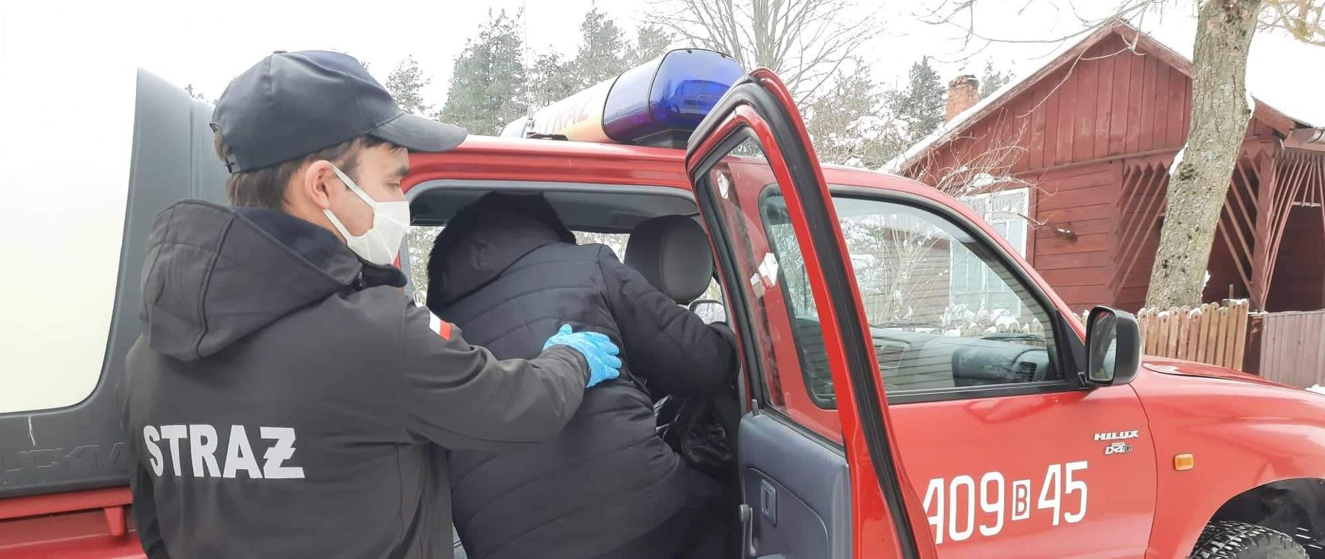 Na zdjęciu widać samochód pożarniczy oraz strażaka pomagającego wsiąść do wozu dla kobiety.