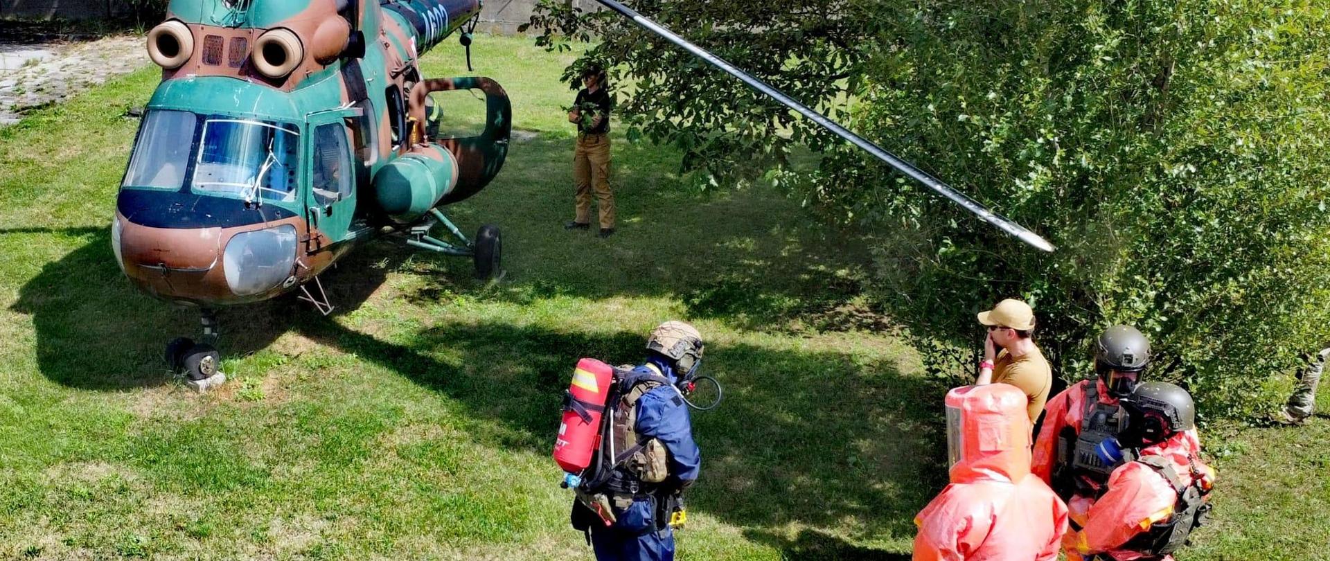 Poligon Centralnej Szkoły PSP, w głębi po lewej śmigłowiec, po prawej czterech uczestników ćwiczeń w kombinezonach do ratownictwa chemicznego oraz instruktor
