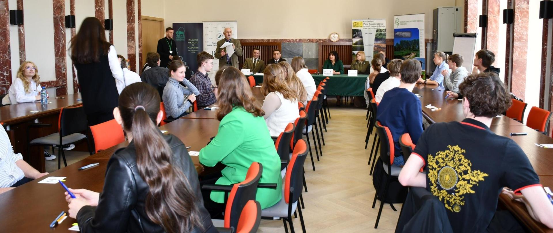 W sali stoły ustawione w długie rzędy, przy nich młodzież, na końcu stół prezydialny, z którym siedzą ludzie, na ścianie godło.